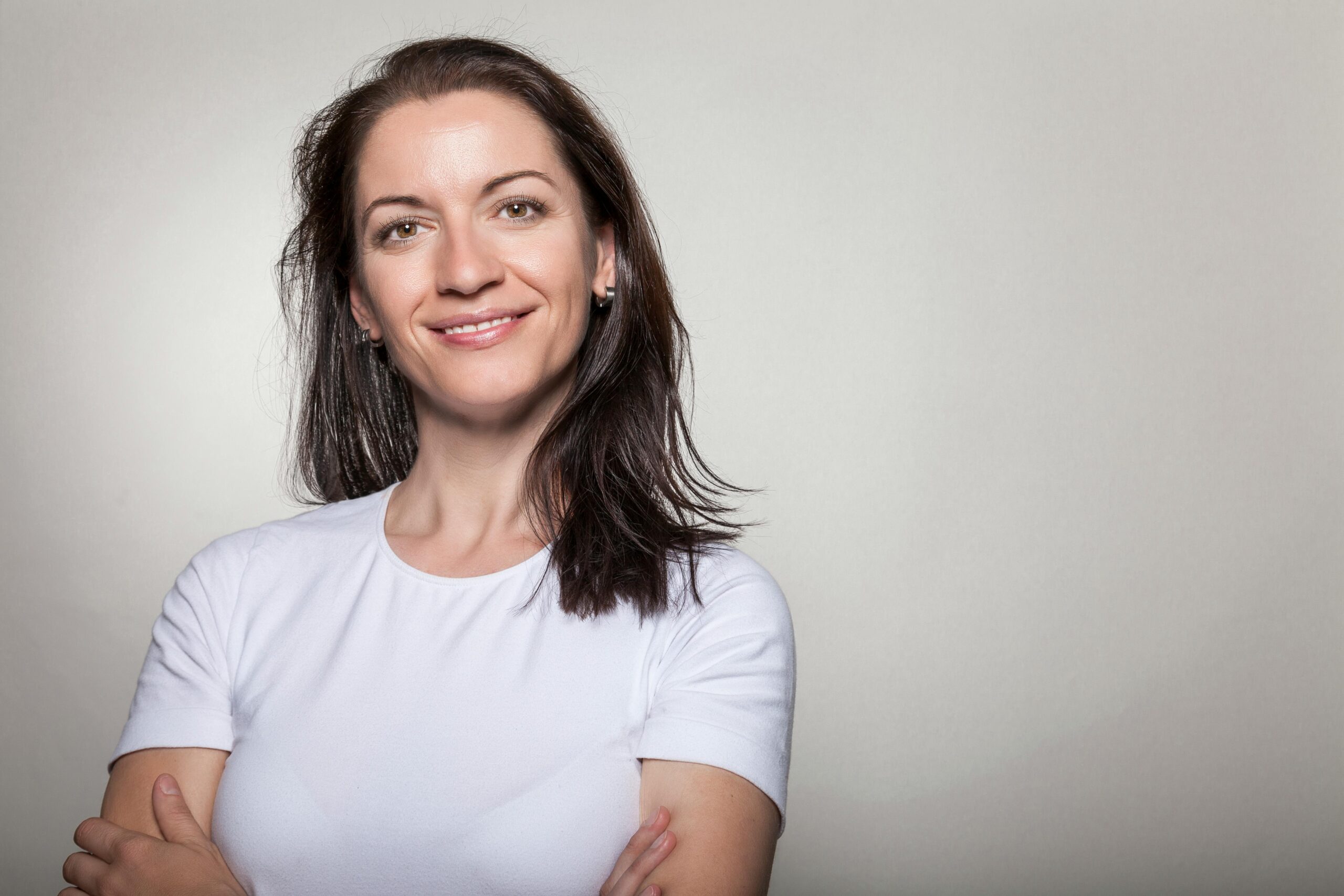 Portrait of a confident woman smiling with arms crossed, wearing a white t-shirt on a gray background.